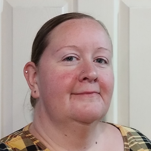 Portrait of a woman with light skin and brown hair tied back, wearing a plaid shirt, standing in front of a white door.