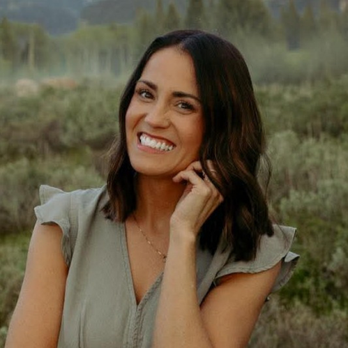Woman with shoulder-length dark hair smiling outdoors in a natural field setting.